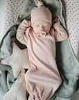 A sleeping baby in a pale pink gown and matching hat lies on a soft green mushie Star Lovey Blanket with a pacifier, while a cream-colored star-shaped pillow rests beside them.