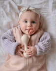 A baby in a light pink outfit and gray knit sweater lies on a white blanket, wearing a headband and holding a beige rattle, using the FRIGG Andersen Fairytale Silicone Pacifier 2-Pack by FRIGG, made safely in Denmark.