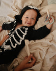 A smiling baby lies on a bed, wrapped in a mushie Organic Cotton Muslin Swaddle Blanket, wearing a black skeleton costume with a hood. The baby holds a teething toy while an adult’s hands gently hold the baby and a wooden utensil.