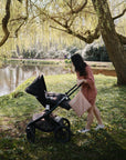 A woman in a pink dress strolls by a pond with her baby, her mushie Water Resistant Wet Bag keeping the diaper bag organized and dry beside willow trees on a sunny day.