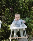 A baby sits outdoors in a high chair, wearing the mushie Long Sleeve Bib in blue and white checks, holding a spoon near their mouth with a bowl and cup on the tray. Green bushes and a white chair are nearby.