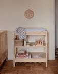 A light wooden changing table with a blue mat holds baby supplies like the mushie Organic Cotton Muslin Swaddle Blanket and toys, next to a crib. Above is a round wall decor with a smiling face. The room features parquet flooring and light walls.