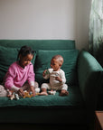 Two young children sit on a green couch. The older child, in a pink sweater, plays with puzzle pieces, while the younger child in white holds the mushie Mix and Match Ice Cream Toy and looks at their sibling. Plants are by the window.