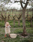 A young child in a light dress stands under a blossoming tree, holding a toy and a FRIGG Lucky Symmetrical Silicone Pacifier (6-18 Months), with pruned trees and a green field in the background.