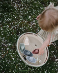 A toddler plays with mushie Bath Boats on grass and flower petals.