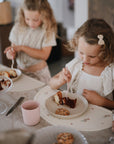 Two girls eat cake with mushie pink Dinnerware Cups and plates at the table.