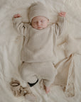 A baby in a beige knit sweater, pants, and hat sleeps on a cream mushie Star Lovey Blanket with arms raised. Nearby are a star-shaped pillow, beige cloth, booties, and a small toy.