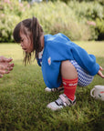 A young toddler in a blue sweater and red socks squats on grass, watching an adult’s hand. Beside her is a mushie Snack Cup—a small, spill-proof silicone container—and a teapot with playful animal designs.