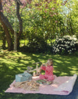 A young girl sits on a picnic blanket beside a mushie Diaper Caddy in a sunny garden.