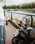 An adult pushes a stroller with a baby holding the mushie Snack Cup. Walking by a riverside with trees and buildings in the background, it’s perfect for toddlers on the go.