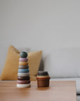 Mushie Stacking Cups Toy on a table, with a sofa and yellow pillow in the background.