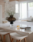 Bright, modern kitchen with a white island, two wooden stools, a brown glass vase with greenery, a small yellow accent, a gray box, and the mushie No-Drop Sun Teether (Sunshine) on display. Large window fills the space with natural light.
