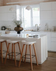 Modern white kitchen with an island and three wooden stools, minimal decor, a large vase with greenery, pendant lights above, and a mushie Lunchbox with built-in compartments placed by the window.