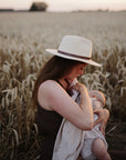 Woman in a hat nursing with mushie Organic Cotton Muslin Swaddle Blanket at sunset in wheat field.