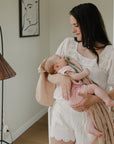 Woman in white holding a baby wrapped in mushie Organic Cotton Muslin Swaddle Blanket, cozy room.