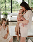 A young girl explores with mushie Kaleidoscopes as a woman holds a baby in a bright room.