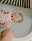 A baby in a striped onesie smiles in a crib with a mushie Extra Soft Muslin Crib Sheet.