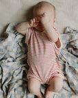Baby lies on mushie Organic Cotton Muslin Swaddle Blanket, hands covering face.