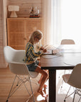 A young girl with long blonde hair and glasses sits at a sunlit dining table, writing in a notebook. Beside her is the mushie Reusable Water Color Pad, an eco-friendly art supply, illuminated by natural light.