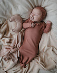 A baby in a rust gown and bonnet sleeps on a cream blanket, holding a pacifier beside the mushie Moon Lovey Blanket—an organic cotton, crescent-shaped security blanket.