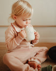 A young child in light pink pajamas sits on the floor, sipping from a white cup with a cute animal design—a perfect scene for enjoying the mushie Bluey Gift Set.