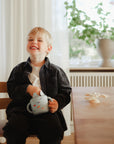 A young boy smiles at a table, holding a light blue teapot. Beside him is the mushie Snack Cup, a spill-proof silicone cup, among small objects. Sunlight pours through white-curtained windows as he enjoys snack time.