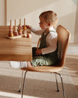 A young child in green overalls plays with a wooden stacking toy at a table, while a mushie Linen Pacifier Clip 2-Pack rests nearby, complemented by warm sunlight filling the room.