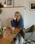 A blonde woman in a blue sweater sits at a wooden table in soft light, sipping from a white cup. The table holds mugs, candles, plates, and a mushie Snack Cup—a spill-proof silicone snack cup for toddlers.