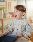 A young child with brown hair smiles excitedly while sitting indoors at a table with a blue tray, wearing the mushie Long Sleeve Bib in a colorful pattern with wide pockets.