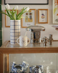 A glass display cabinet holds blue and white teacups, a vase of tulips, a Mushie Bluey x Mushie Silicone Training Cup + Straw, and metallic salt and pepper shakers. Framed art hangs on the wall behind.