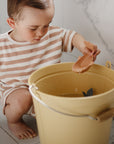 A child plays with Mushie Whale Bath Stackers, scooping water from a yellow bucket.