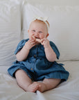 Smiling baby in blue dress holding mushie Figurine Teether while sitting on a bed.