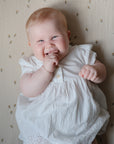 Smiling baby on mushie Extra Soft Muslin Crib Sheet, hand near mouth, wearing white dress.