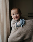 A smiling baby in a beige knit sweater, held by an adult and clutching the mushie No-Drop Dino Teether (Tradewinds), is bathed in soft natural light streaming through a nearby curtained window.