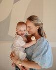 A woman in a blue puff-sleeve top holds a smiling baby dressed in white, who is grasping the mushie Silicone Baby Rattle Toy. They are in front of a neutral-toned abstract background, sharing a joyful sensory moment.
