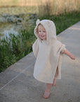 A toddler wearing the mushie Bear Poncho Towel, made from organic cotton and featuring bear ears, walks barefoot along a paved path by a pond and tall grass.