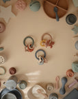Various pastel mushie baby toys and feeding items, such as the Space Teething Ring made from food-grade silicone, plates, cups, spoons, and stacking toys for sensory development, are neatly arranged in a circle on a beige surface.