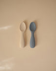 Two mushie Silicone Toddler Starter Spoons, one cream and one blue, rest side by side on a light beige surface with soft natural light from the left—ideal for encouraging independent feeding.