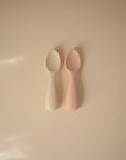 Two mushie Silicone Toddler Starter Spoons, one off-white and one light pink, rest side by side on a beige surface. Both have the brand name embossed on their handles, ideal for encouraging independent feeding. Soft light casts gentle shadows nearby.