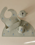The mushie Bluey x Mushie Silicone Placemat, pale green silicone baby bib, and two cups with Bluey-inspired cartoon dogs and white stars are arranged neatly on a light beige surface.