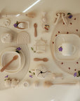 Overhead view of mushie Square Dinnerware Plates for children in beige BPA-free plastic, with cups, spoons, toys, and teethers arranged on two placemats. Purple flowers and petals are scattered among the items.