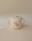 The mushie Snack Cup is a cream-colored, spill-proof silicone cup with two handles, a soft lid, and a pink floral design—ideal for toddlers and shown on a plain beige background.