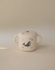 The mushie Snack Cup is a cream-colored, spill-proof silicone cup with two handles and lid, featuring a simple whale and fish design—ideal for toddlers and shown on a plain, light background.