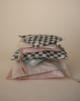 A stack of pastel mushie Water Resistant Wet Bags, including two with blue and white checkerboard patterns, displayed against a beige background. Each bag features a zipper, diaper bag storage design, and a small white label.