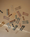 A selection of mushie Dinnerware Fork and Spoon Sets in brown, blue, green, yellow, and pink silicone and plastic are displayed in a scattered pattern on a beige surface; all sets are dishwasher-safe.