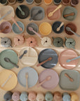 Mushie’s pastel silicone children’s tableware—including bowls, plates, cups, utensils, and a spill-proof Snack Cup—is neatly arranged in rows on a light surface, showcasing their soft minimalist design.