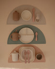 Three sets of mushie Silicone Toddler Starter Spoons with matching silicone plates, cups, utensils, and bowls are arranged on semicircular placemats in beige, green, and pink against a tan background.
