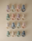Sixteen Mushie Silicone Fresh Food Feeders, in soft pastel blue, pink, green, and cream, are arranged neatly in a four-by-four grid on a beige background—safe for easy self-feeding and made from food-grade silicone.
