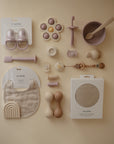 Assorted beige and pastel baby items, including the mushie Silicone Baby Rattle Toy, bib, teethers, pacifiers, a feeding set, and food-grade silicone utensils are neatly arranged on a neutral background.