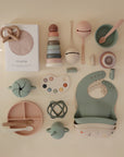 A flat lay of pastel baby items—bibs, plates, bowls, toys, stacking rings, a mushie Snack Cup (spill-proof silicone), utensils, and a muslin cloth—are neatly arranged on a cream background.